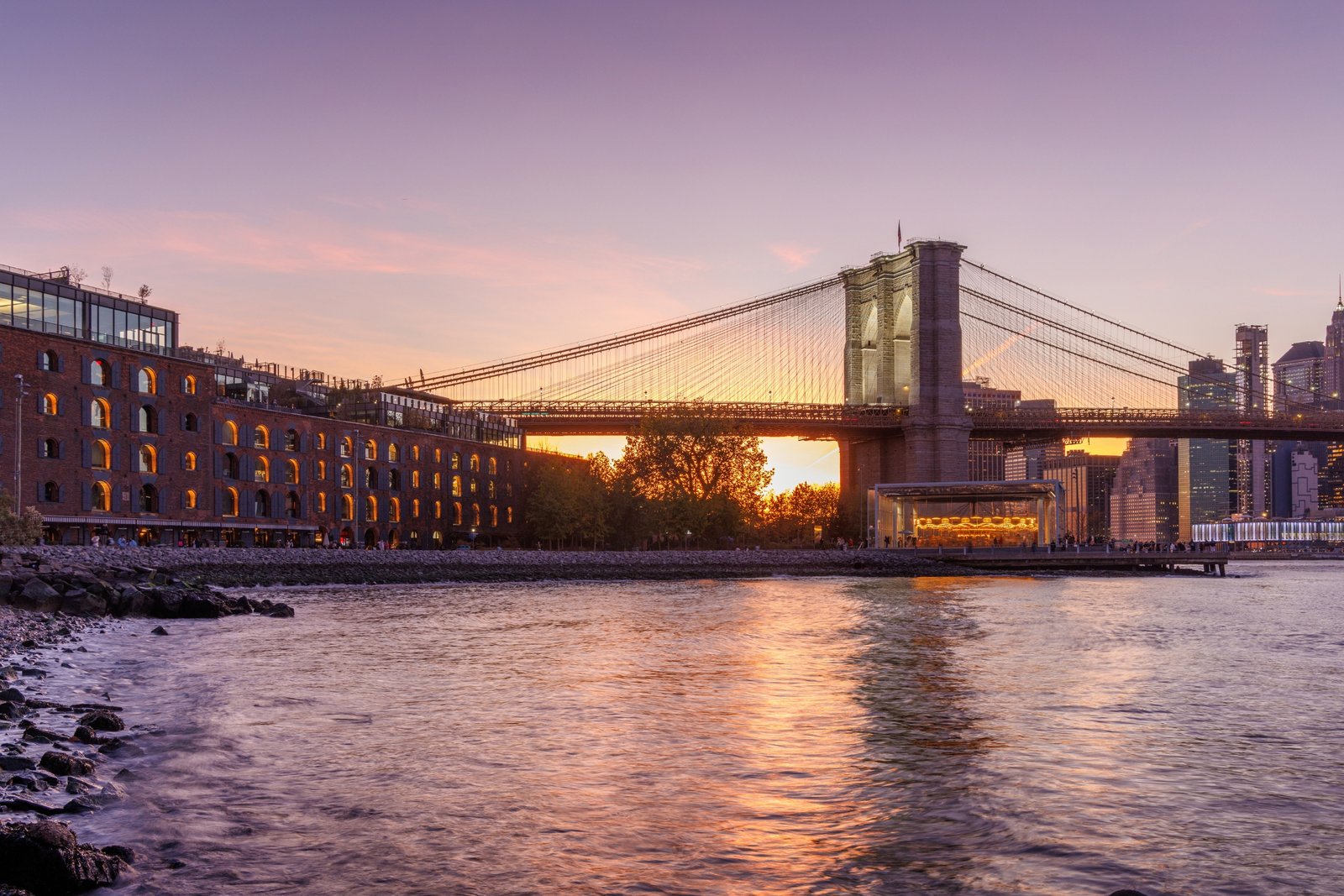 A panoramic sunset view of the Brooklyn Bridge stretching towards Manhattan, with skyscrapers illuminated in warm hues against the fading sky