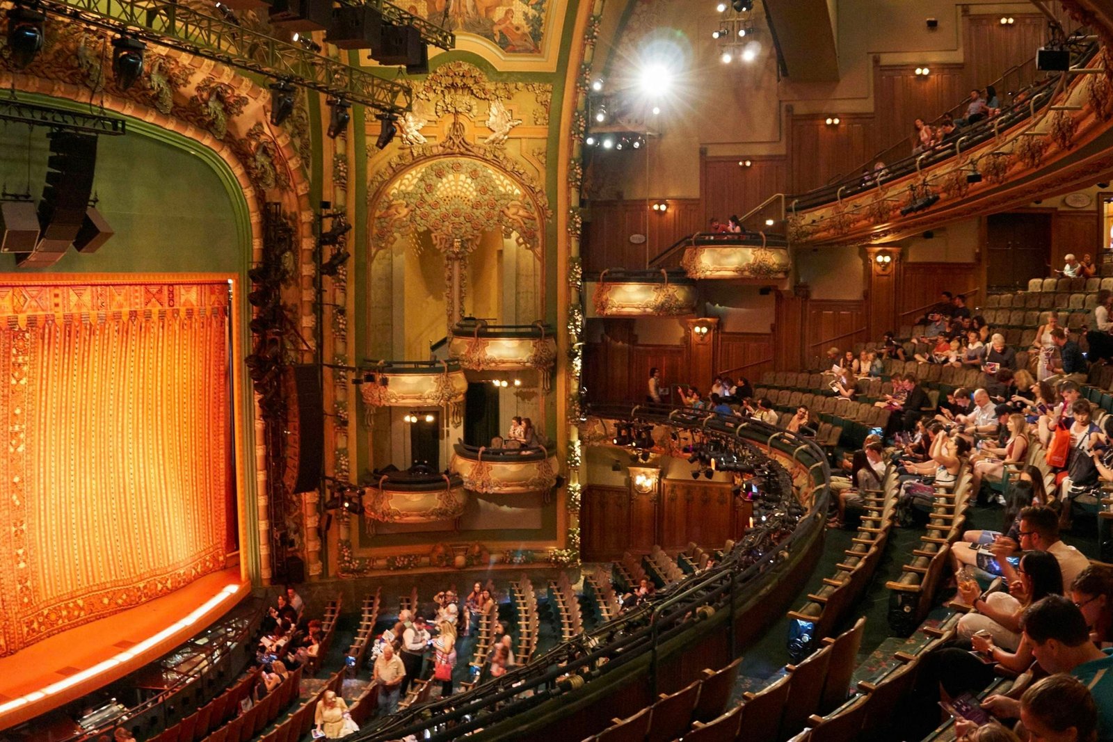 Audience seated inside an ornate Broadway theater with a vibrant red and gold curtain on stage, waiting for the performance to begin. The interior features intricate architectural details, balcony boxes, and warm lighting.