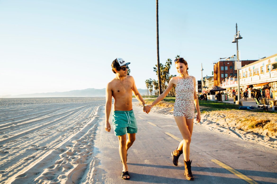 Young couple walking hand in hand along a sunny beach boardwalk, with the ocean and palm trees in the background.